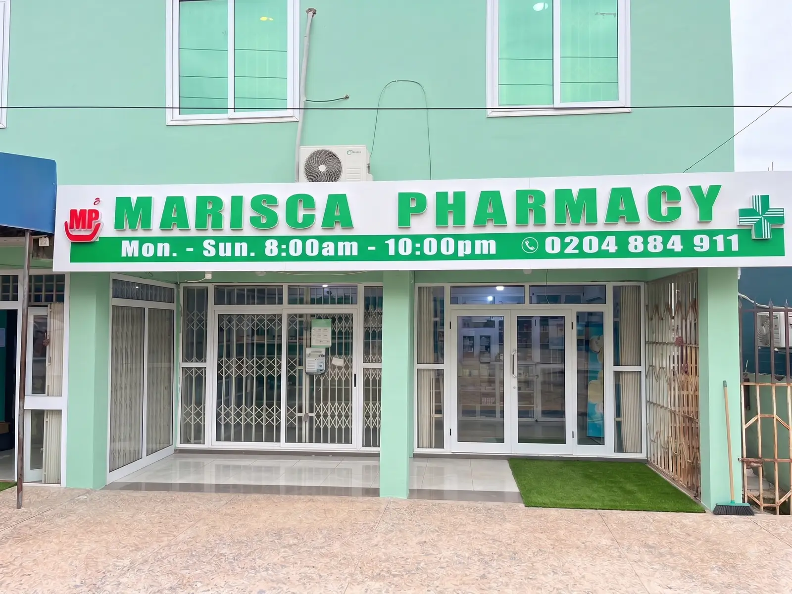 The Accra branch of Marisca Pharmacy, a mint green building with a white sign reading Marisca Pharmacy, open Monday to Sunday 8am to 10pm.
